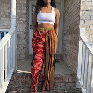 May include: A white cropped tank top paired with high-waisted cargo pants featuring a vibrant red and gold geometric pattern. The pants have a drawstring waistband, wide legs, and black rectangular side pockets on each thigh area.