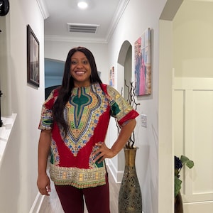 Pode incluir: Uma camisa dashiki de manga curta com uma base vermelha vibrante e padrões geométricos intrincados em amarelo, verde e azul ao redor do decote e da bainha, combinada com leggings sólidas de cor vermelho-escura até aos tornozelos.