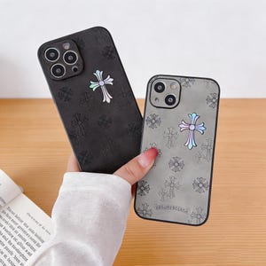 Puede incluir: Dos fundas para teléfono con diseños de cruces. Una funda es negra con una cruz iridiscente grande y cruces más pequeñas. La otra funda es gris con una cruz iridiscente grande y cruces más pequeñas. Las fundas se sostienen en una mano.