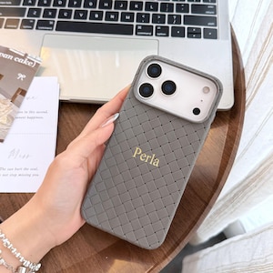 Könnte beinhalten: Graue Handyhülle mit geflochtener Textur, mit dem Wort "Perla" in goldfarbenen Buchstaben. Die Hülle hat einen Kameraausschnitt und wird in der Nähe eines Laptops und einer Holzoberfläche gehalten.