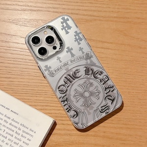 Op de afbeelding: Transparante telefoonhoes met een zilver Chrome Hearts ontwerp. De hoes heeft een herhalend kruispatroon en het logo van het merk. De telefoonhoes ligt op een houten oppervlak naast een boek.