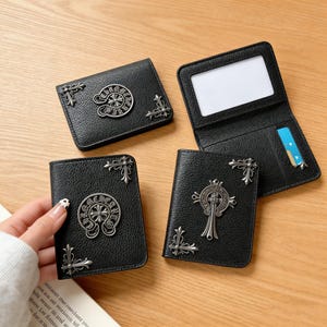 Puede incluir: Carteras de cuero negro con adornos plateados. Las carteras presentan cruces metálicas decorativas y detalles florales en las esquinas. Una cartera está abierta, revelando una ventana de identificación transparente y una ranura para tarjetas. Sobre una superficie de madera.