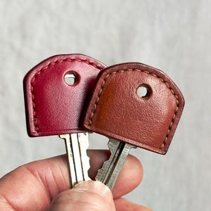 May include: Two key covers made of leather, one burgundy and one brown, are held in a hand. The covers have a stitched edge and a hole for the key ring. The keys are silver.