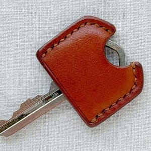 May include: A key with a silver blade is partially inserted into a brown leather key holder. The key holder is rectangular with rounded corners and stitched edges. The leather has a reddish-brown hue.