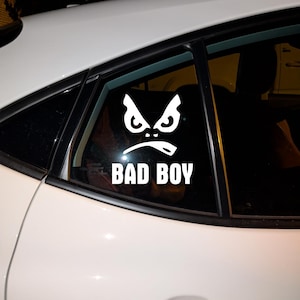 May include: White vinyl decal of a stylized angry face with the text "BAD BOY" below it. The decal is applied to the rear window of a white car. The design features bold lines and a menacing expression.