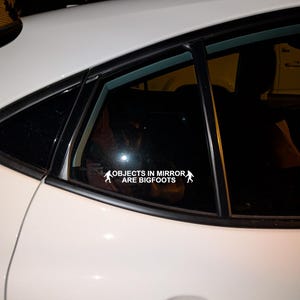 May include: White car window sticker with the text "OBJECTS IN MIRROR ARE BIGFOOTS" and two Bigfoot silhouettes. The sticker is applied to the rear window of a white car, with the car's body visible.
