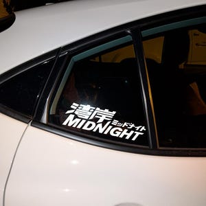 May include: White vinyl decal with Japanese characters and the word "MIDNIGHT" in a bold, sans-serif font. The decal is affixed to the window of a white car, with the car's interior visible through the glass.