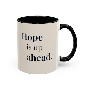 May include: A ceramic coffee mug with a beige exterior and a black interior and handle. The mug features the words "Hope is up ahead" in navy blue lettering.