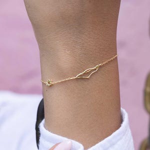 May include: A delicate gold bracelet featuring a star-shaped charm and a stylized outline of a landmass. The bracelet is worn on a wrist, showcasing its fine chain and minimalist design. The jewelry is set against a soft pink background.