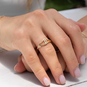 May include: A gold-colored ring with a floral design. The ring features an openwork band with a small flower-like detail at the center. The ring is worn on a finger, showcasing its delicate and elegant style.