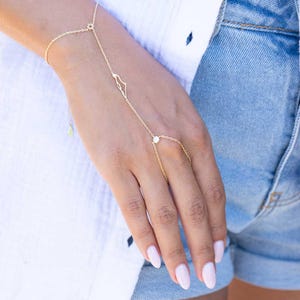 May include: A delicate gold hand chain bracelet. The bracelet features a starburst design on the wrist, a teardrop charm, and a small gem connecting to the middle finger. The hand chain is worn on a person's hand.
