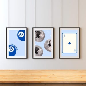 Puede incluir: Tres impresiones artísticas enmarcadas sobre un fondo azul claro. Las impresiones presentan una bola ocho azul con el texto "Lucky Girl", tres bolas de discoteca y un as de picas. Los marcos son negros.