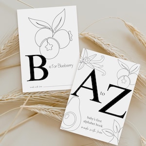 May include: Two white rectangular cards with black text and line art illustrations. One card features a large letter B, the text B is for Blueberry, and a blueberry drawing. The other card displays A to Z, baby's first alphabet book, and fruit art.