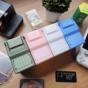 以下が含まれることがあります： 緑、ピンク、白、青の4色の3Dプリントされたカードホルダー。ホルダーは、ヒンジ付きの蓋が付いた長方形のデザインです。 他のアイテムには、ポラロイドカメラ、小さな植物、トランプの箱が含まれます。