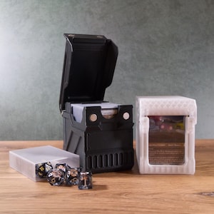 May include: A black card storage box with an open lid, a clear plastic dice case, and a white card case. The black box is open, revealing cards inside. Several dice are visible in the clear case. The white case is also open, showing a card.