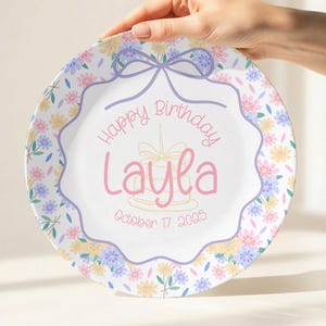 Könnte beinhalten: Ein weißer Teller mit einem floralen Rand in Pastellfarben. Der Teller trägt den Text "Happy Birthday Layla" mit dem Datum "October 17, 2025". Eine Schleife und eine Kuchendarstellung sind ebenfalls enthalten.
