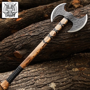 May include: A double-bit axe featuring a textured black metal head with sharpened silver edges. The handle is crafted from light brown wood with a prominent grain, wrapped in black leather cord lacing and a solid black grip at the base.