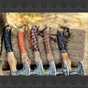 May include: A collection of six Viking axes with wooden handles and intricately designed metal blades. The handles are wrapped in various colors of leather, including black, brown, and a checkered pattern. The axes are displayed on a wooden log.