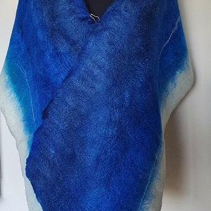 May include: A blue felted shawl with a gradient effect, transitioning from deep blue to white. The shawl is draped over a black mannequin and secured with a silver pin. The bottom right corner of the image has the text "KNITONEFELTTWO".