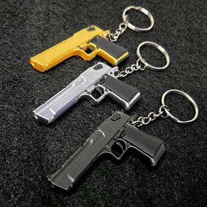 May include: Three metal pistol-shaped keychains, each featuring a split ring attached by a short chain. The keychains are finished in metallic gold, silver, and matte black, with black grip detailing on the handle of each miniature firearm replica.