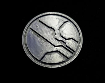 May include: A circular silver-toned medallion featuring a raised geometric pattern with recessed lines. The surface displays a textured finish with visible layered details, creating a distinct relief design within the round frame.