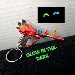 May include: A red ray gun keychain with a black handle, blue accents, and a yellow sight. The keychain features a glow-in-the-dark element, shown in an inset box with green and blue light. The text GLOW IN THE DARK is printed in green.
