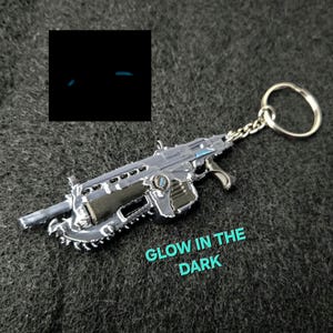 May include: A silver-colored miniature gun keychain with a chain and ring. The text "GLOW IN THE DARK" is visible. The image also shows a small square with two glowing blue lines.