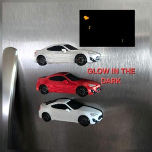 May include: Three sports car magnets arranged vertically, featuring two white cars and one red car, all with black wheels and windows. A black rectangular inset shows glowing yellow lights, and the red text reads GLOW IN THE DARK.
