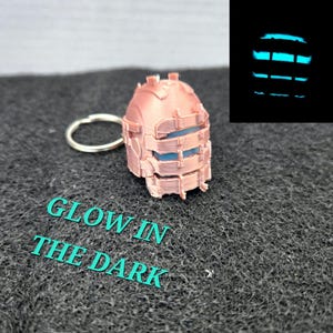 May include: A copper-colored, 3D-printed keychain shaped like a helmet with blue accents. The keychain has a silver ring. The text "GLOW IN THE DARK" is printed in teal. A smaller image shows the helmet glowing blue.