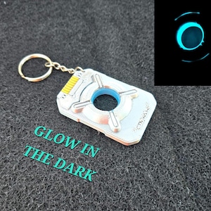 May include: A silver-colored keychain with a blue center and a gold-colored keyring. The keychain has a unique design with four arms extending from the center. The text "GLOW IN THE DARK" is visible. A separate image shows the keychain glowing blue.