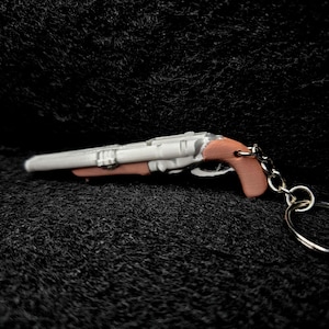 May include: A miniature, detailed double-barreled shotgun keychain. The gun features a white body with brown grip and trigger, attached to a silver keyring. The item is set against a dark, textured background.
