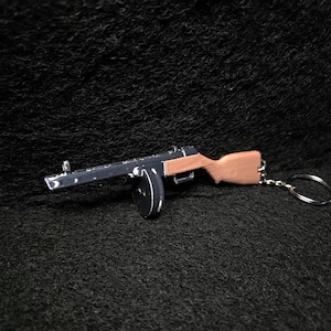 May include: Miniature black and brown toy machine gun keychain. The keychain features a detailed replica of a firearm, with a wooden stock and a circular magazine. The gun is attached to a silver keyring.