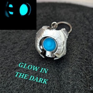 May include: A silver-colored pendant with a bright blue orb at its center. The pendant has a detailed, geometric design and a small loop at the top for a chain. The text "GLOW IN THE DARK" is visible below the pendant. A separate image shows the orb glowing.