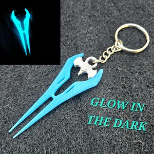 May include: A light blue keychain featuring a glowing, stylized weapon design. The keychain includes a silver-colored chain and ring. The text "GLOW IN THE DARK" is visible. The image also shows a glowing version of the weapon.