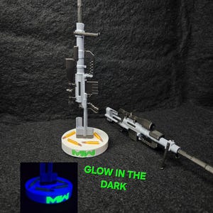 May include: A gray and black toy rifle with a scope and a stand. The stand is white with the letters "MW" in green. The text "GLOW IN THE DARK" is also in green.