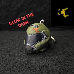 May include: A green and black helmet keychain with a silver keyring. The helmet has orange accents and the text "GLOW IN THE DARK" in red. A small, glowing yellow shape is in the upper right corner.