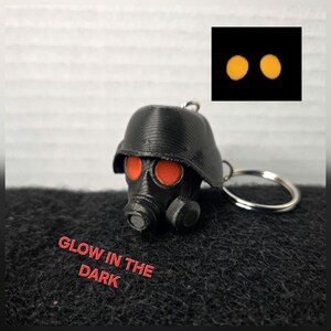May include: Black gas mask keychain with red eye lenses. The keychain has a silver ring and the text "GLOW IN THE DARK" in red. A small inset shows two glowing orange circles.