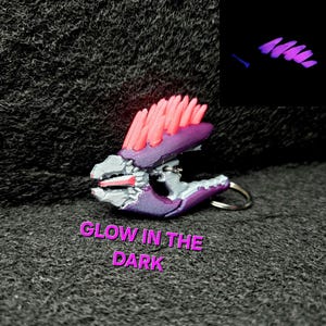 May include: A purple and gray keychain with a design that glows in the dark. The keychain has a red and pink accent. The text "GLOW IN THE DARK" is displayed in pink. A small inset shows the glowing effect.