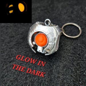 May include: A silver-colored keychain with an orange center and a key ring. The keychain has a geometric design and the words "GLOW IN THE DARK" in red. A smaller inset image shows the keychain glowing.