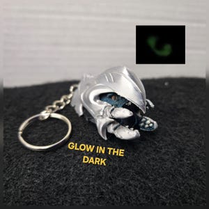 May include: A silver-colored keychain featuring a creature with a metallic shell and blue textured skin. The text GLOW IN THE DARK is printed in yellow, and a small inset shows the creature emitting a green glow in a dark environment.