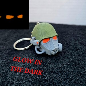 May include: A gray and green helmet-shaped keychain with orange accents and a silver keyring. The text "GLOW IN THE DARK" is printed in red. The helmet's eye lenses and other details glow in the dark.