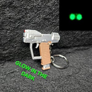 May include: A silver and brown miniature gun keychain with a red accent. The keychain has a silver ring and a brown handle. The text "GLOW IN THE DARK" is in green. The image also shows two glowing green circles.