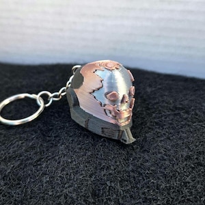 May include: A 3D-printed keychain featuring a stylized skull design. The skull is primarily silver with bronze and black accents, attached to a silver keyring. The keychain is resting on a dark, textured surface.
