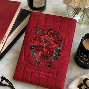 May include: A rectangular, quilted red fabric notebook cover featuring an embroidered design of a central anatomical heart surrounded by roses and skulls. The text "People & Paperwork New York" is embroidered in red thread near the bottom edge.