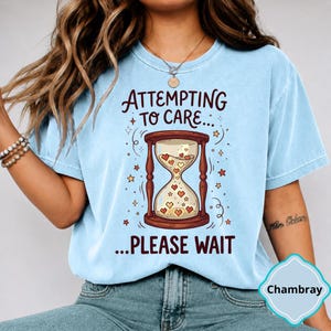 May include: A light blue short-sleeve t-shirt featuring a graphic of a brown hourglass filled with small hearts. The text above the hourglass reads "ATTEMPTING TO CARE..." and the text below reads "...PLEASE WAIT" in dark brown lettering.
