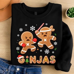 May include: Black sweatshirt featuring a gingerbread man boxing and another holding a candy cane. The word "GINJAS" is written in festive lettering with a Santa hat and Christmas tree. Snowflakes and green sprigs complete the design.