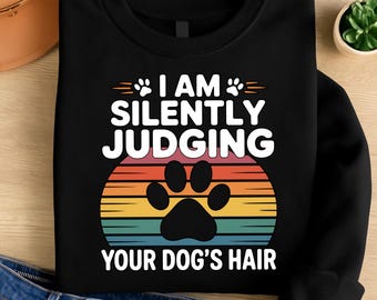 May include: Black sweatshirt with the text "I AM SILENTLY JUDGING YOUR DOG'S HAIR" in white. The design includes a paw print and a colorful sunset graphic. A fun, casual top for dog lovers.