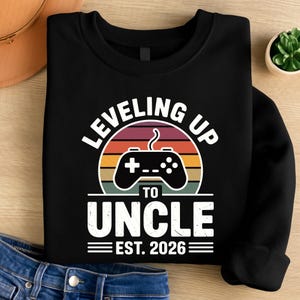 May include: Black sweatshirt with a graphic that reads "LEVELING UP TO UNCLE EST. 2026." The graphic features a retro sunset design with a video game controller in the center. The sweatshirt is folded, with a pair of blue jeans visible in the bottom left corner.