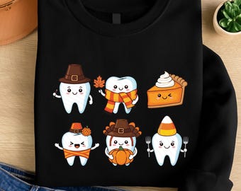 May include: Black sweatshirt featuring six cartoon teeth dressed in Thanksgiving-themed attire. One tooth wears a pilgrim hat, another holds a leaf, and a third holds a slice of pumpkin pie. The other teeth are holding a pumpkin, corn, and a fork.