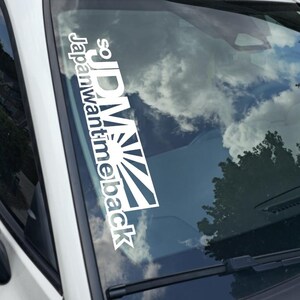 May include: White vinyl decal with the text "so JDM Japan want time back" and a rising sun design. The decal is affixed to the windshield of a white car, with a reflection of the sky and trees.